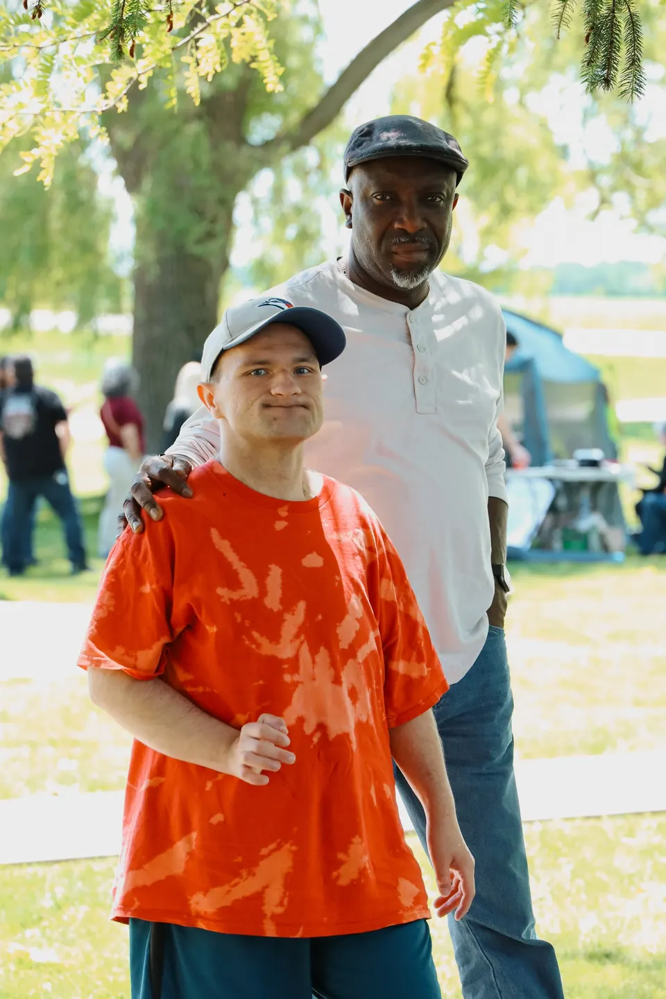 A young man in an orange t-shirt and cap smiling outdoors at a summer event, a hand resting on his shoulder