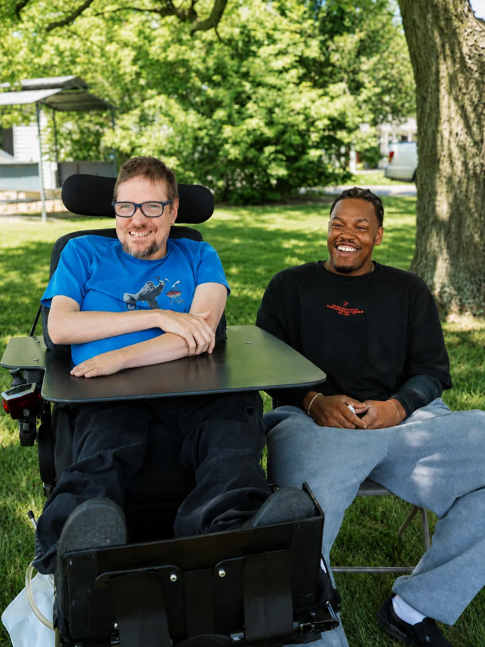A man in a power wheelchair and a younger man sitting together on the grass under a tree, both smiling at the camera