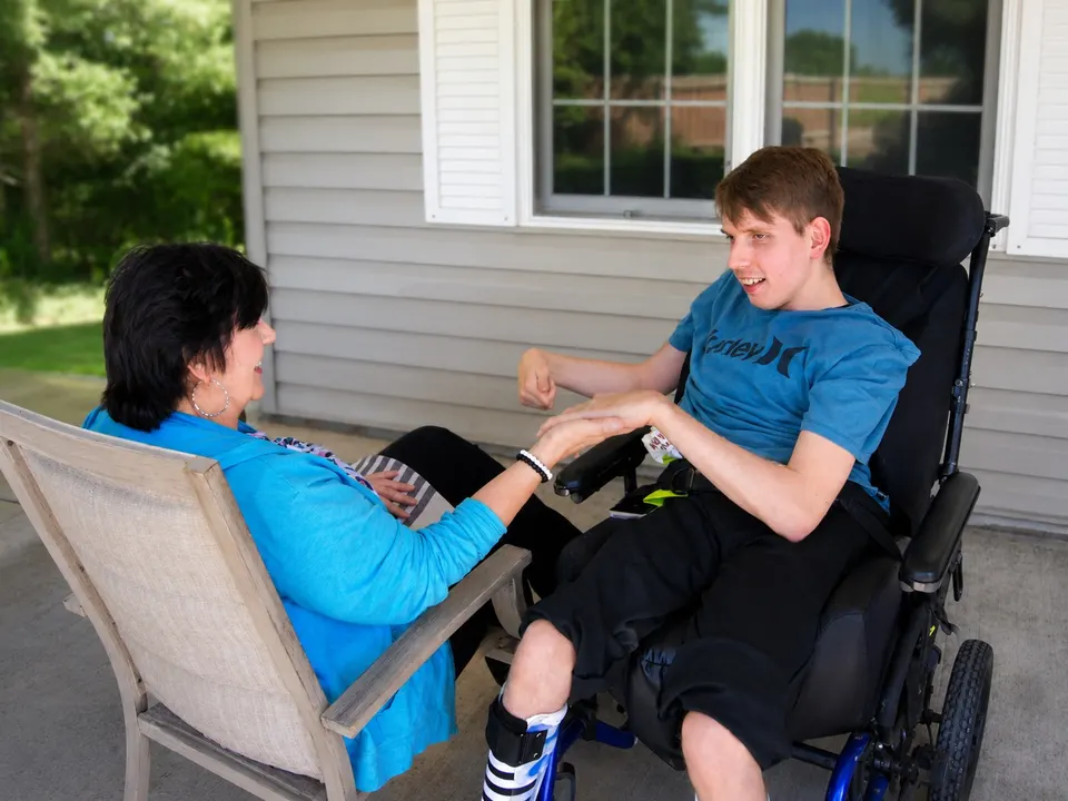 A young man in a power wheelchair laughing and gesturing mid-conversation, with a woman sitting beside him on the lawn