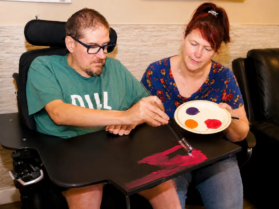 A man in a wheelchair painting on canvas while a support worker holds the palette