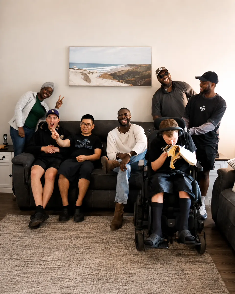 Seven friends laughing together in the living room of a Hopewell home, some on the couch and one in a power wheelchair