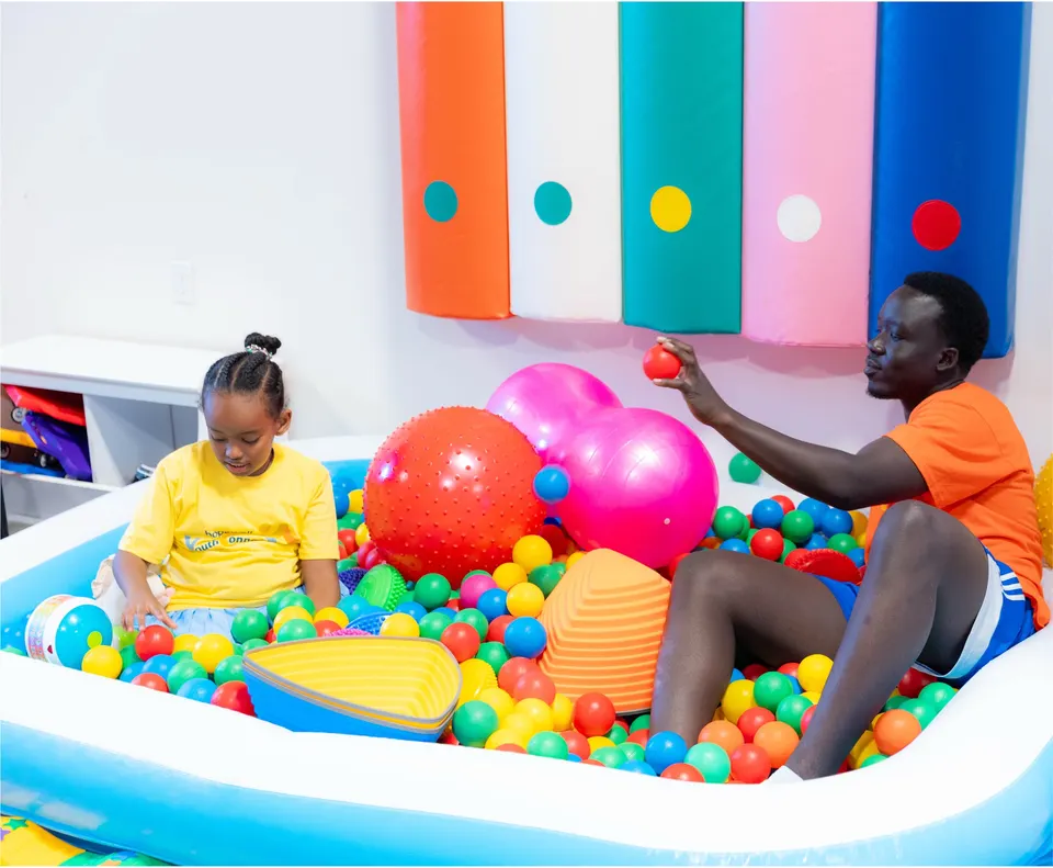 A support worker and a child playing together in a ball pit full of colourful balls at a Hopewell program.