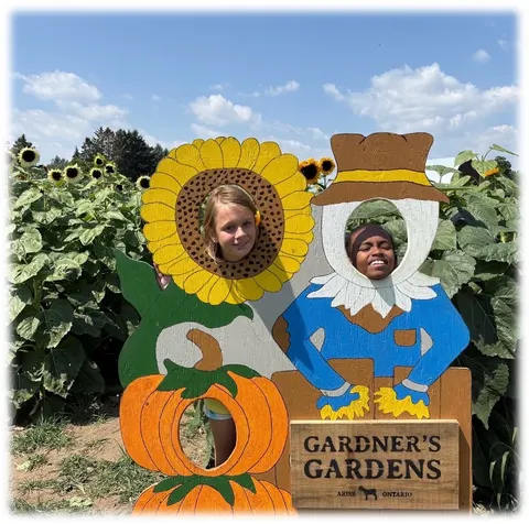 Two children smiling through the cut-out holes of a painted sunflower and scarecrow photo board on a farm outing.
