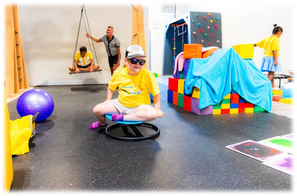 A girl in a Hopewell Youth Connect t-shirt spinning on a sensory chair in the gym while other children play on a swing and build with soft blocks behind her.