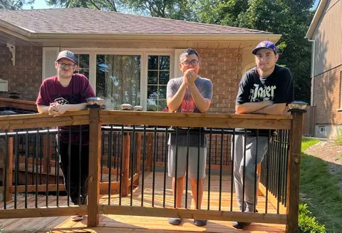 Three housemates standing together at the front deck railing of their home on a sunny day